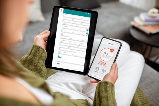 L'immagine rappresenta l'uso della **tecnologia digitale nella sanità**, mostrando una persona che utilizza un **tablet** e uno **smartphone** per gestire la propria salute da casa.  <br /><br />Sul **tablet** è visibile una **cartella clinica digitale** o un **modulo di registrazione sanitaria**, con campi dettagliati per l'inserimento di dati personali e informazioni mediche. Il layout ordinato e il design con intestazione **verde petrolio** suggeriscono un software di **gestione sanitaria online**.  <br /><br />Sul **telefono**, è aperta un'app di **monitoraggio della frequenza cardiaca**. Il display mostra un battito cardiaco di **92 BPM** e un avviso di **aritmia anomala**, consigliando di contattare un medico. L'interfaccia pulita e intuitiva evidenzia l'importanza della **telemedicina e del controllo remoto della salute**.  <br /><br />L'ambiente circostante è accogliente, con **luce naturale** e un tavolino con libri e una tazza, trasmettendo un senso di relax mentre l'utente si prende cura della propria salute con strumenti digitali.  <br /><br />Questa immagine è ideale per comunicare concetti chiave come **sanità digitale, telemedicina, monitoraggio della salute, cartelle cliniche elettroniche e tecnologia per il benessere**, fondamentali nel settore della **salute digitale e del medical software**.