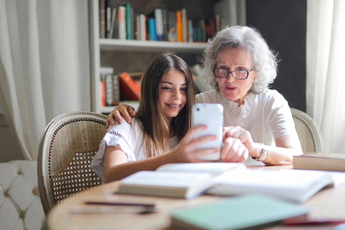 L'immagine mostra una **nonna e sua nipote** che usano insieme uno **smartphone** in un ambiente accogliente con libri e quaderni sul tavolo. La scena rappresenta il **legame intergenerazionale**, l'**apprendimento digitale per gli anziani** e l'uso della **tecnologia per connettere le generazioni**.  <br /><br />### **Parole chiave SEO:**  <br />**Nonna e nipote con smartphone, digitalizzazione anziani, connessione tra generazioni, apprendimento tecnologico per senior, famiglia e tecnologia, alfabetizzazione digitale per anziani, insegnare la tecnologia agli anziani.**